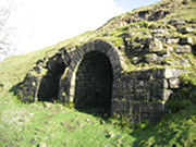 Two adjacent stone-built limekilns, each with a circular arch at the top, built into a grass bank